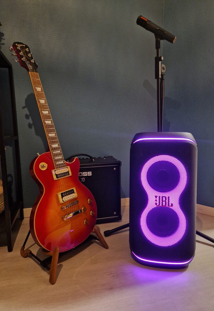 Kuvassa seinää vasten oranssi sähkökitara, musta kitaravahvistin, mustat mikkiteline ja mikki ja musta JBL-kaiutin, jossa violetti valo etuosassa.